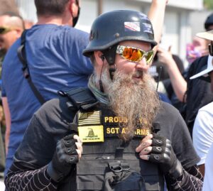 A bearded man wears a black motorcycle helmet and a ballistic vest with patches reading "Proud Boys" and "Sgt At Arms", as well as a black and yellow American Flag and a Gadsden Flag patch. 