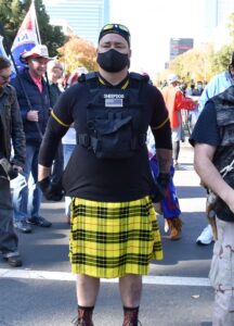 A man wears a black and yellow "Fred Perry"-style polo shirt and a black and yellow kilt. Over the tio, he wears a black ballistic vest with the word "Sheep Dog" and a backwards American Flag. He has tattoos visible on both forearms. 