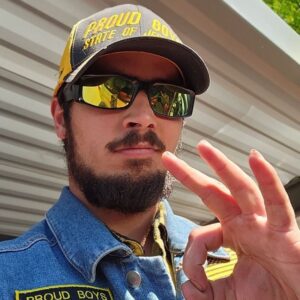 A man with a dark beard gives the "OK" sign with his hand. His yellow and black hat reads "Proud Boys Satate of Jefferson" and a black and yellow patch on his denim jacket reads "Proud Boys"