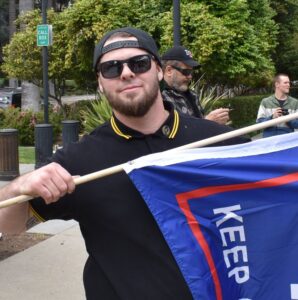 A photo of a bearded man in a black and yellow "Fred Perry"-Style polo shirt and sunglasses, holding a Trump 2020 flag. 