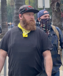 A Photo of a heavyset man with a red-brown beard. the Outline of a ballistic vest is visible beneath his black T-shirt. He wears a yellow bandana and ablack and red hat. . A tattoo is visible running down his right interior forearm
