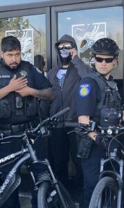 A m a man wearing a gray hoodie, a skull mask, sunglasses, and a white shirt with the German war eagle visible on it holds his phone up to his ear. He's surrounded by bicycle cops from Sacramento Police Department.