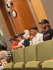 five men sit in a row of seats in a city council chamber. On the right side is Jeffrey Perrine, with a short bear and mullet. he is wearing a hat with the meme Honkler on it and a shirt reading "Pureblood"