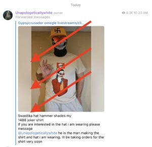 A screenshot from Telegram. Unapologeticallywhite shares a post from "Gypsycrusader omegle livestreams/clips". The post reads: swastika hat hammer shades my 1488 Joker shirt. if you are interested in the hat I am wearing please message @unapologeticallywhite he is making the shirt and hat I am wearing. I'll be taking orders for the shirt soon. The attatched Photo shows a man standing indoors. He is wearing a skull mass, a yellow hat with a black swastika, and a T-shirt featuring a man and a joker outfit holding a gun with a flag coming out of the end. The flag has the number 1488 on it. The man holds his hand in an OK gesture. 