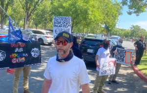 Jeff Perine, wearing sunglasses and a hat with the letters FAFO on it stands in front of a group of masked men wearing signs.