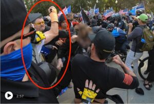 A group of men in Proud Boys attire have fistss out attacking a single person, whose face is blurred. On hand, which is circled and raised above the others, has an orange cast on it. 
