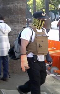 A man walks by the camera, wearing a Sacramento Proud Boys hat, a black and yellow gaiter, a tan ballistic vest, and a gray backpack. He has an orange cast on one hand and a black cast on the other. 