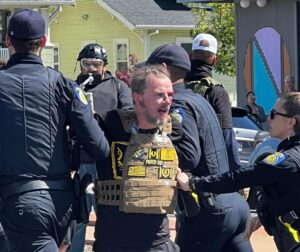 A photo of police leading away Jonathan Giusto, wearing a tan ballistic vest with an assortment of Proud Boys patches on it, and bleeding from above his left eye. 
