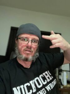 A man with glasses and a scraggly, graying beard holds his middle finger up to the camera. 